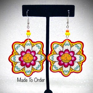 May include: A pair of colorful, handmade earrings featuring a white flower with a jack-o'-lantern face in the center. The earrings are made of wood and have a red, yellow, and white color scheme. "Made To Order" is written below the earrings.