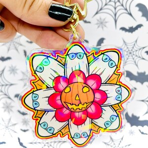 May include: A clear, holographic keychain featuring a flower design with a jack-o'-lantern center and ghost petals wearing glasses. The keychain has a gold star-shaped clasp.