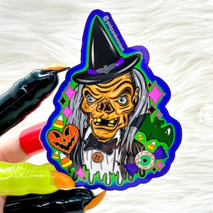 May include: A sticker featuring a cartoon witch with green and purple accents. The witch is wearing a black hat with a purple band and has a green and purple dripping effect around her. The sticker also features a pumpkin, candy corn, a bat, and an eyeball.