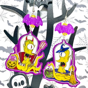 Puede incluir: Un par de pendientes de temática de Halloween con un amuleto de murciélago morado y un extraterrestre amarillo con un solo ojo, uno sosteniendo un tridente y el otro una escoba, ambos con un caldero y una calabaza.
