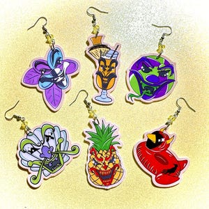 May include: A collection of six novelty earrings with various cartoon character designs. The earrings feature images of a flower, a cocktail, a planet, a shell, a pineapple, and a devil. Each earring has a silver hook and yellow bead accents.