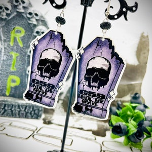 Può includere: Un paio di orecchini a forma di bara di colore viola e nero con un teschio e il testo "669 Under the Grave".