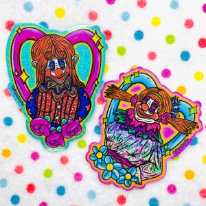 May include: Two glitter stickers featuring creepy clown characters with exaggerated features and bright colors. The stickers are shaped like hearts with a sparkly blue background.