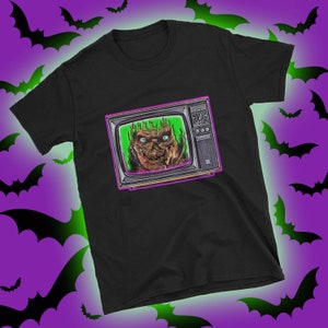 May include: Black t-shirt featuring a graphic of a monster's face emerging from a vintage television set. The television is surrounded by a purple glow.