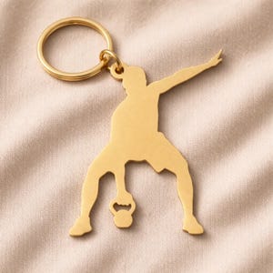 May include: A gold-coloured metal keyring featuring a silhouette of a person lifting a kettlebell. The keyring has a circular ring at the top for attaching keys. The person is depicted in a dynamic pose, with one arm extended and the other holding the kettlebell.