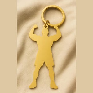 May include: A gold-colored keychain featuring a stylized silhouette of a muscular person flexing their biceps. The keychain has a circular ring for attaching keys and is set against a beige fabric background.