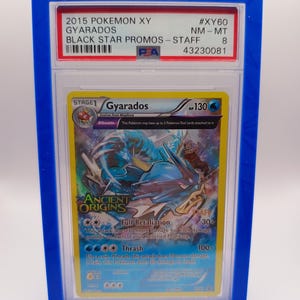 May include: A PSA graded Pokemon card in a blue plastic case. The card is a Gyarados from the Ancient Origins set. The card is graded NM-MT 8 and has the number #XY60. The card is a Black Star Promo - Staff card.