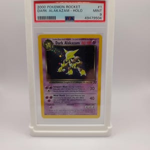 May include: A graded Pokemon card featuring Dark Alakazam from the Rocket set. The card is holographic and in mint condition. The card is graded a 9 by PSA and has a unique number 49479504.