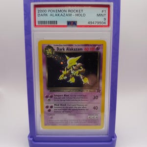 May include: A graded Pokemon card in a protective case. The card is a holographic Dark Alakazam from the Rocket set. The card is in mint condition and has a grade of 9. The card's number is #1. The card's text reads "Evolves from Dark Kadabra. Put Dark Alakazam on the Stage 1 card. Dark Alakazam 60 HP.  ... Teleport Blast You may switch Dark Alakazam with 1 of your Benched Pok&eacute;mon. (Do the damage before switching the Pok&eacute;mon.) Mind Shock Don't apply Weakness and Resistance for this attack. (Any other effects that would happen after applying Weakness and Resistance all happen.)"