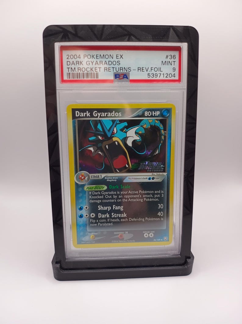 May include: A graded Pokemon card in a protective case. The card is a Dark Gyarados from the 2004 Pokemon EX: Team Rocket Returns set. The card is in mint condition and has a 9 out of 10 rating. The card features a dark blue and red Gyarados with a dark blue background. The card text reads "Dark Scale" and "Dark Streak".