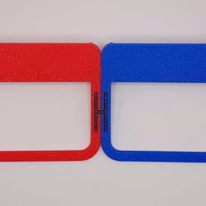 May include: Two plastic frames, one red and one blue, with a rectangular opening in the centre. The frames are textured and have a slightly raised edge.