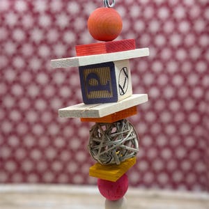 Puede incluir: Un colorido juguete para pájaros suspendido de una cadena metálica. El juguete presenta un bloque de madera con la letra "P" y un diseño de fútbol americano, bloques de madera, una bola tejida y varios elementos de madera y esféricos en tonos naranja, rojo, amarillo, rosa y blanco.