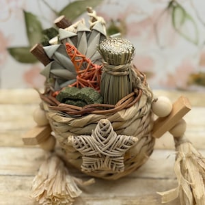 May include: A woven seagrass basket filled with natural materials including a small orange rattan star, a bundle of dried grass, green compressed hay, and wooden sticks. The basket features a woven star on the front and dangling raffia tassels.