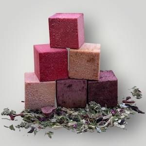 Puede incluir: Una pirámide de siete cubos texturizados de colores, en tonos rosa, rojo y marrón. Los cubos están apilados uno encima del otro, con una dispersión de hierbas y hojas secas en la base. Los cubos parecen estar hechos de un material sólido.