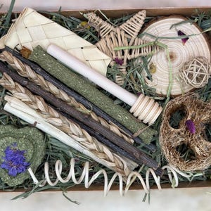 May include: A selection of natural pet toys and treats presented in a box. The contents include woven shapes, wooden sticks, a wooden log slice, a heart-shaped treat with a blue flower, and a loofah, all on a bed of green hay.