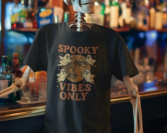 May include: Black t-shirt with the text "SPOOKY VIBES ONLY" in orange, featuring a moon and ghost graphic. The shirt is worn by a skeleton. The background is a bar setting.