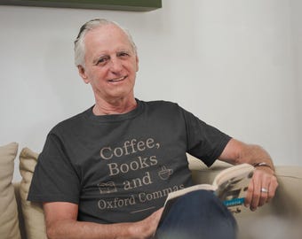 May include: A dark gray t-shirt with the text "Coffee, Books and Oxford Commas" in a light brown color. The shirt features a book and coffee cup graphic. The person is sitting on a beige couch and reading a book.