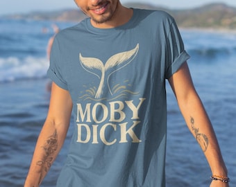May include: A blue t-shirt with the text "MOBY DICK" and a whale tail design. The shirt is being worn by a person standing near the ocean. The design is in a light beige color.