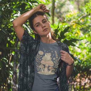 May include: A gray t-shirt featuring a whimsical illustration of a frog sitting on a mushroom, surrounded by flowers and plants. The shirt is worn with a dark patterned shawl. The background is a lush green forest.