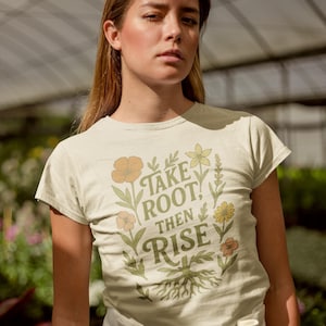 May include: Cream-colored t-shirt with the text "Take Root, Then Rise" in green and gold. The design includes floral and root illustrations. The shirt is short-sleeved and has a crew neck.