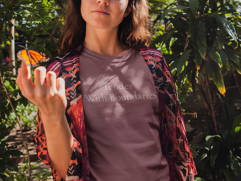 May include: A mauve t-shirt with the text "Tender With Boundaries" is worn under a patterned kimono. A monarch butterfly rests on a hand. The background features lush green foliage, creating a natural setting.