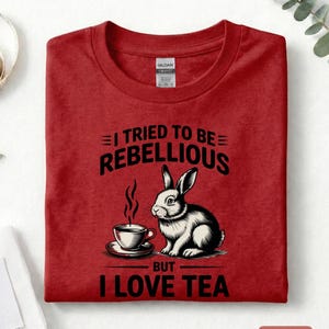 Puede incluir: Camiseta roja con el texto "I TRIED TO BE REBELLIOUS BUT I LOVE TEA". El gr&aacute;fico presenta un conejo en blanco y negro con una taza de t&eacute;. La camiseta est&aacute; doblada y tiene una etiqueta.