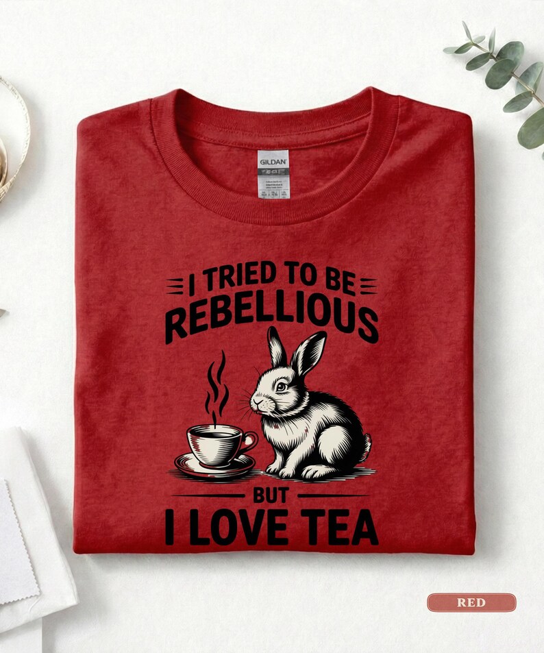 Puede incluir: Camiseta roja con el texto "I TRIED TO BE REBELLIOUS BUT I LOVE TEA". El gr&aacute;fico presenta un conejo en blanco y negro con una taza de t&eacute;. La camiseta est&aacute; doblada y tiene una etiqueta.