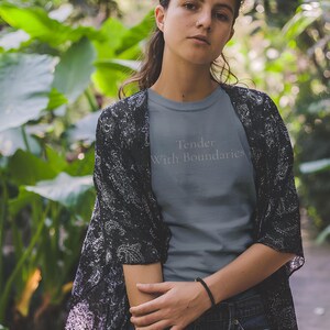 May include: A gray t-shirt with the text "Tender With Boundaries" in light gray. The person is wearing a black and white patterned kimono and dark jeans. The background is a lush green garden. The image is taken outdoors.