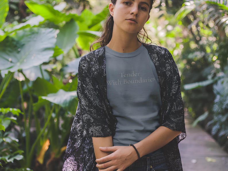 May include: A gray t-shirt with the text "Tender With Boundaries" in light gray. The person is wearing a black and white patterned kimono and dark jeans. The background is a lush green garden. The image is taken outdoors.