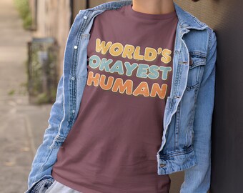 May include: A maroon t-shirt with the text "WORLD'S OKAYEST HUMAN" in yellow, blue, and orange. The person is wearing a light blue denim jacket and gray sweatpants. The person is also wearing sunglasses and holding a white cup.