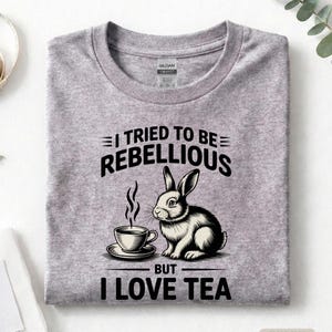Puede incluir: Camiseta gris deportivo con un gr&aacute;fico de un conejo y una taza de t&eacute;. El texto dice "I TRIED TO BE REBELLIOUS BUT I LOVE TEA". La camiseta est&aacute; doblada.