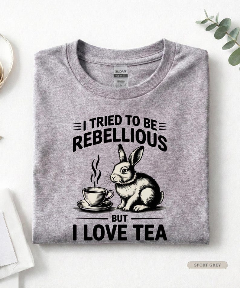 Puede incluir: Camiseta gris deportivo con un gr&aacute;fico de un conejo y una taza de t&eacute;. El texto dice "I TRIED TO BE REBELLIOUS BUT I LOVE TEA". La camiseta est&aacute; doblada.