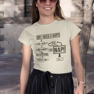 May include: A light beige t-shirt with the text "DO I NEED A NAP?" and a flowchart. The flowchart includes questions like "AM I TIRED?" and "AM I CRANKY?" with arrows leading to "NAP!" and "Zzz...".