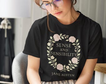 May include: Black t-shirt featuring the text "Sense and Sensibility" within a floral wreath, with "Jane Austen" below. The shirt is worn by a person wearing glasses and holding a book.
