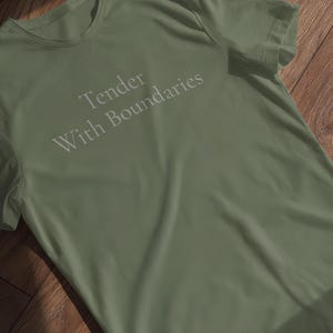 May include: An olive green t-shirt featuring the text "Tender With Boundaries" in light grey. The shirt is made of a soft material and is displayed on a wooden surface.