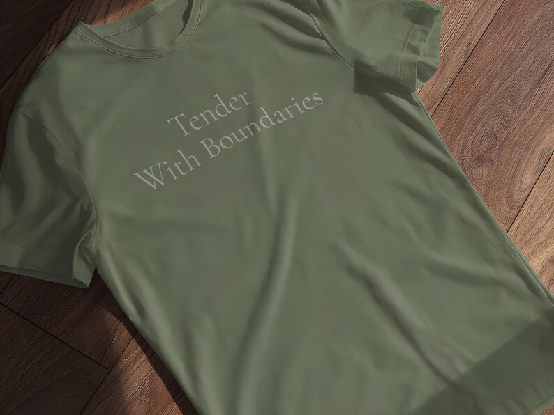 May include: An olive green t-shirt featuring the text "Tender With Boundaries" in light grey. The shirt is made of a soft material and is displayed on a wooden surface.