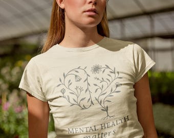 May include: A light beige t-shirt with a floral design in the shape of a brain. The words "MENTAL HEALTH matters" are printed below the design in a gray color. The shirt is short-sleeved.