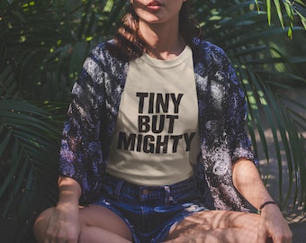 May include: A cream-coloured t-shirt with the text "TINY BUT MIGHTY" in bold black letters. The person is wearing a dark blue patterned kimono and denim shorts. The background features lush green foliage.
