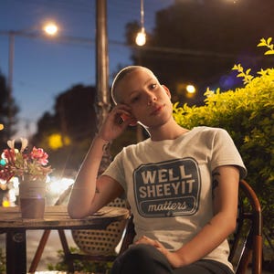 May include: A light-coloured t-shirt with the text "WELL SHEEYIT matters" in a navy blue and white design. The person is sitting at a wooden table with a small vase of flowers, under string lights.