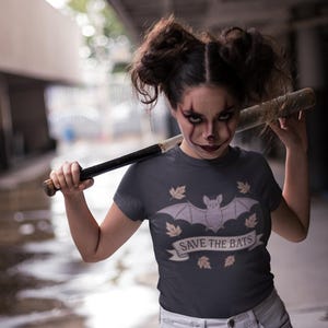 May include: A dark grey t-shirt with a bat graphic and the text "SAVE THE BATS". The person in the image is holding a baseball bat over their shoulder. The person is wearing makeup.