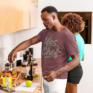 May include: A maroon t-shirt with the text "Yes Chef" in a light gray font. The image shows a person in a kitchen setting, with a basket of fruit, a bottle of wine, and other kitchen items on the counter.