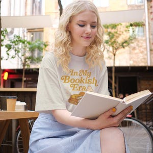 May include: Cream-colored t-shirt with the text "In My Bookish Era" in a retro font, featuring a stack of books. The person is wearing a light blue striped skirt and reading a book with a white cover. The scene is set outdoors.