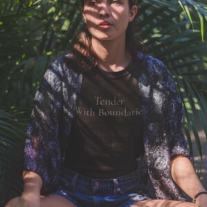 May include: A person wearing a black t-shirt with the text "Tender With Boundaries" and denim shorts, with a patterned kimono. The background features lush green foliage, creating a natural setting.