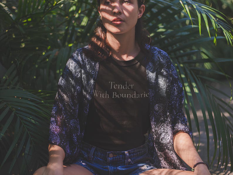 May include: A person wearing a black t-shirt with the text "Tender With Boundaries" and denim shorts, with a patterned kimono. The background features lush green foliage, creating a natural setting.