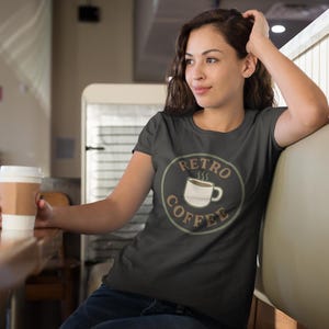 May include: Dark grey t-shirt with a circular "Retro Coffee" design featuring a coffee cup. The person is holding a coffee cup with a brown sleeve. The background includes a booth and a counter.
