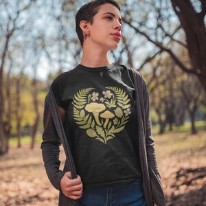 May include: Black t-shirt with a heart-shaped design of mushrooms, ferns, and flowers in shades of green and yellow. The person is wearing blue denim shorts and a gray cardigan. The background is a blurred forest.