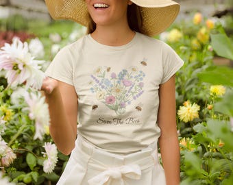 May include: Cream-colored t-shirt featuring a heart-shaped floral design with bees. The design includes various flowers in pastel colors and the text "Save The Bees" in a script font. The shirt is worn outdoors.