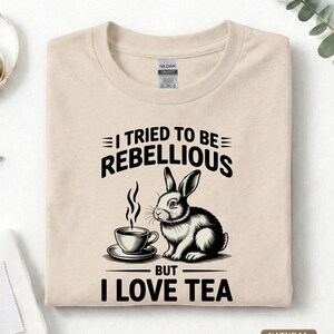 Puede incluir: Camiseta beige con un gr&aacute;fico en blanco y negro de un conejo y una taza de t&eacute;. El texto dice "I TRIED TO BE REBELLIOUS BUT I LOVE TEA". La camiseta est&aacute; hecha de un material suave.