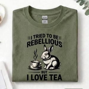 Puede incluir: Camiseta verde militar con el texto "I TRIED TO BE REBELLIOUS BUT I LOVE TEA". Presenta una ilustraci&oacute;n en blanco y negro de un conejo y una taza de t&eacute;. La camiseta est&aacute; doblada.