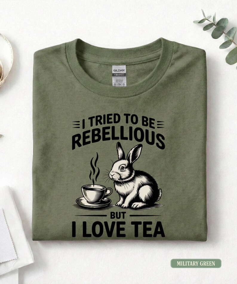 Puede incluir: Camiseta verde militar con el texto "I TRIED TO BE REBELLIOUS BUT I LOVE TEA". Presenta una ilustraci&oacute;n en blanco y negro de un conejo y una taza de t&eacute;. La camiseta est&aacute; doblada.